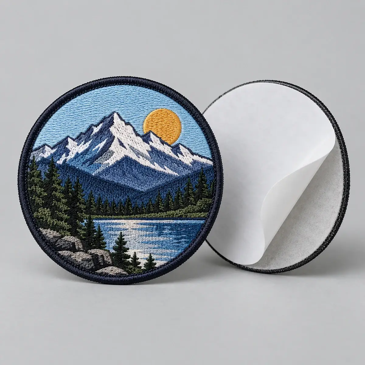 Round embroidered mountain lake patch standing beside its peeled-back adhesive liner.