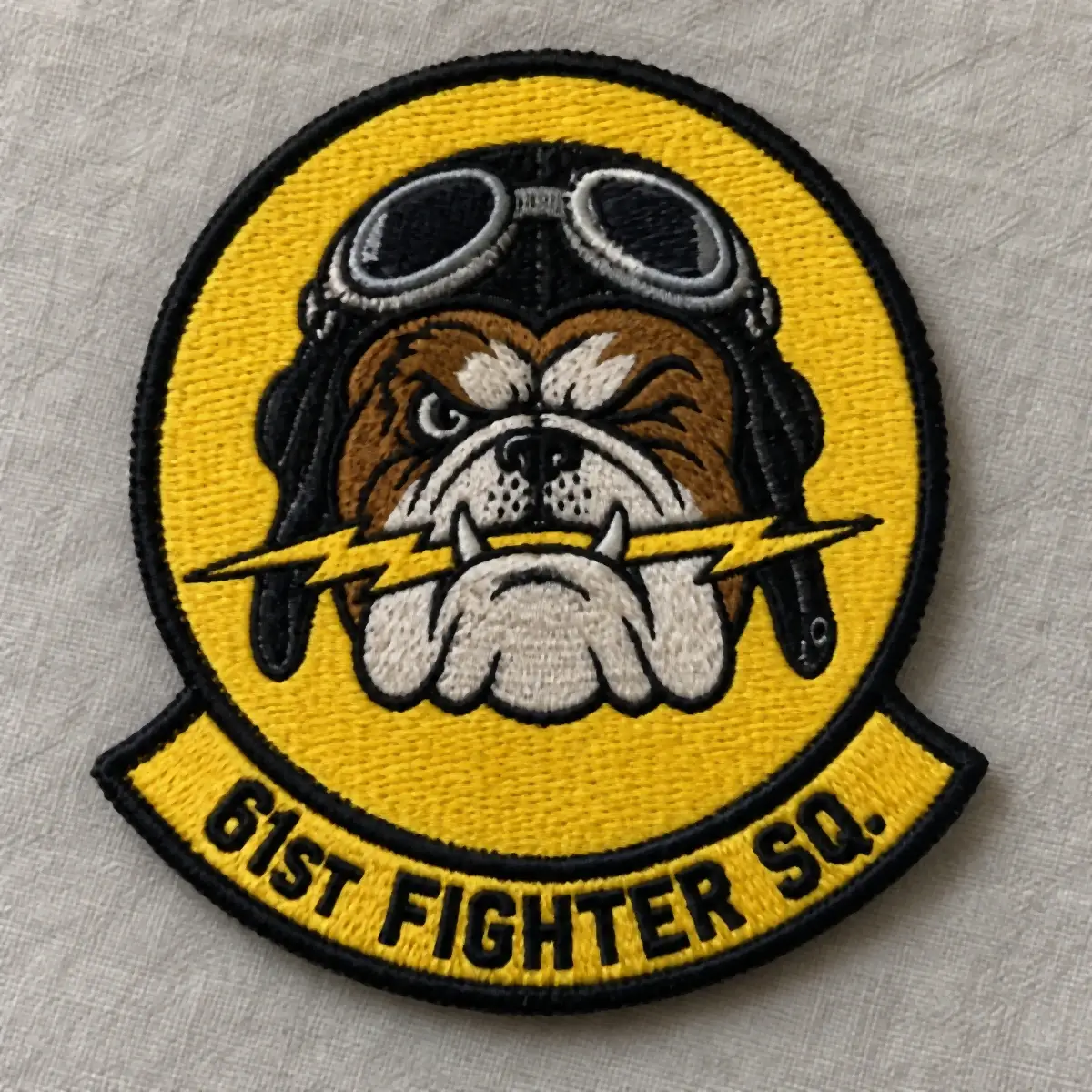Yellow and black embroidered 61st Fighter Sq. patch with a bulldog in a flight helmet biting a lightning bolt.