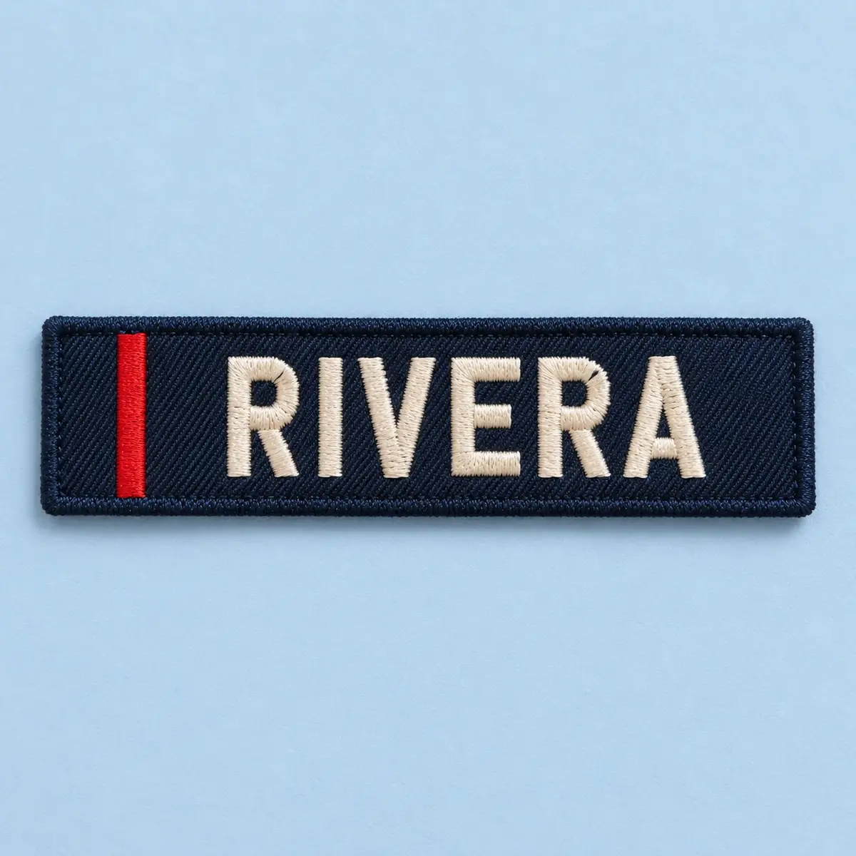 Navy embroidered RIVERA name tag with a red stripe on pale blue.
