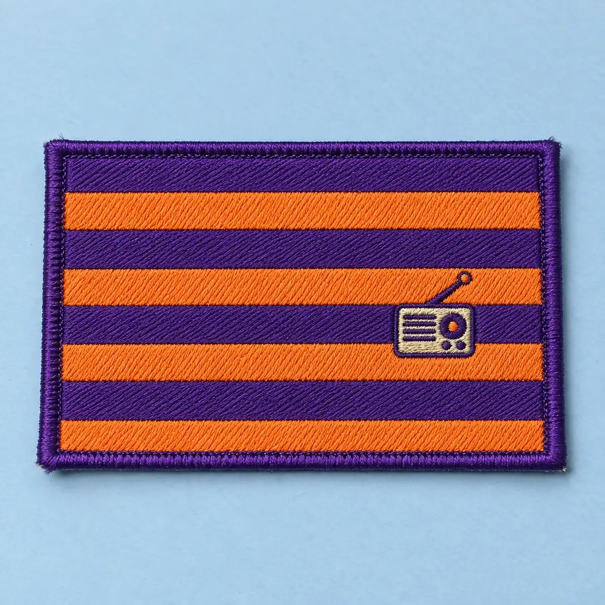 Embroidered purple and orange striped flag patch with a radio emblem, on pale blue.