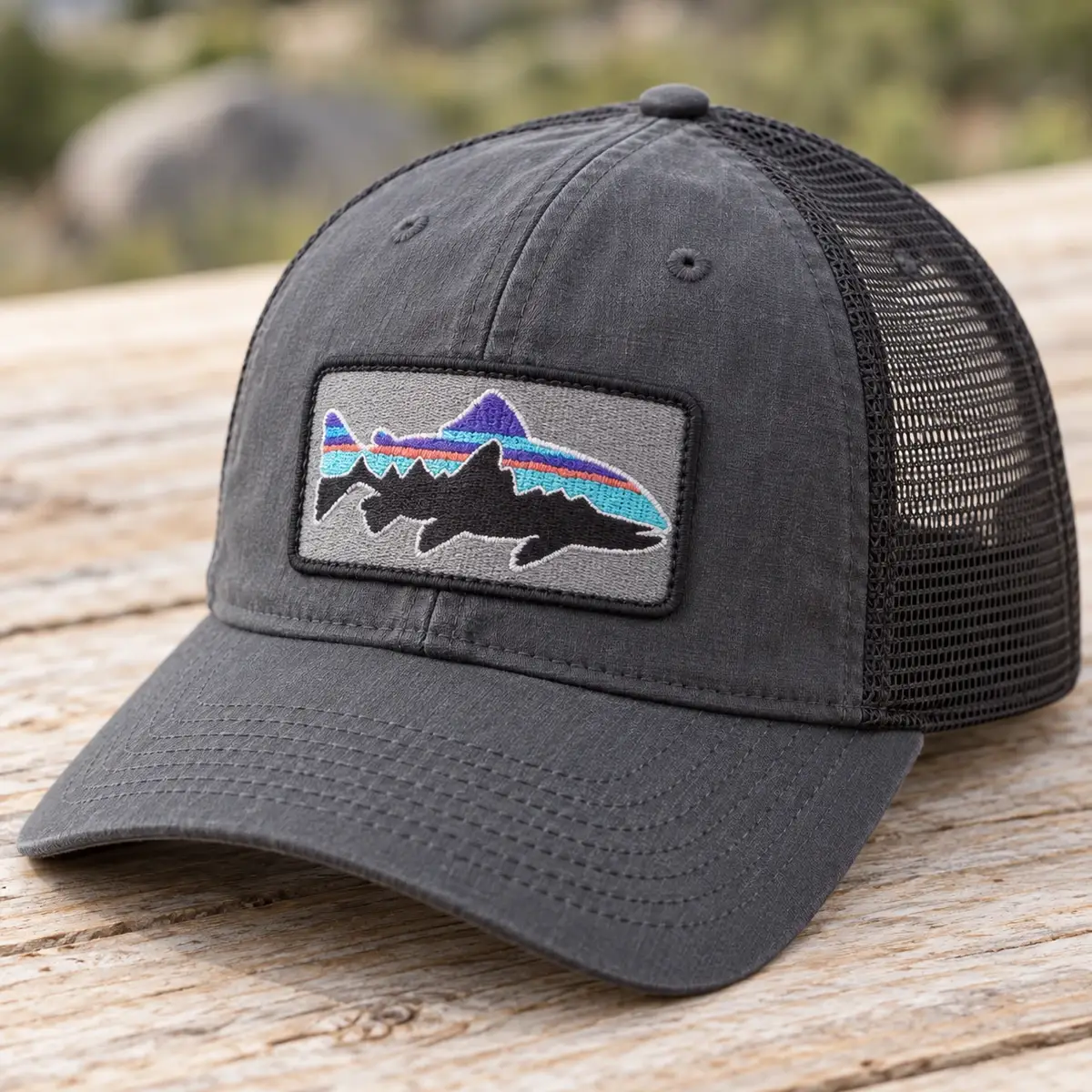 Rectangular embroidered trout and mountains patch on a gray trucker hat.