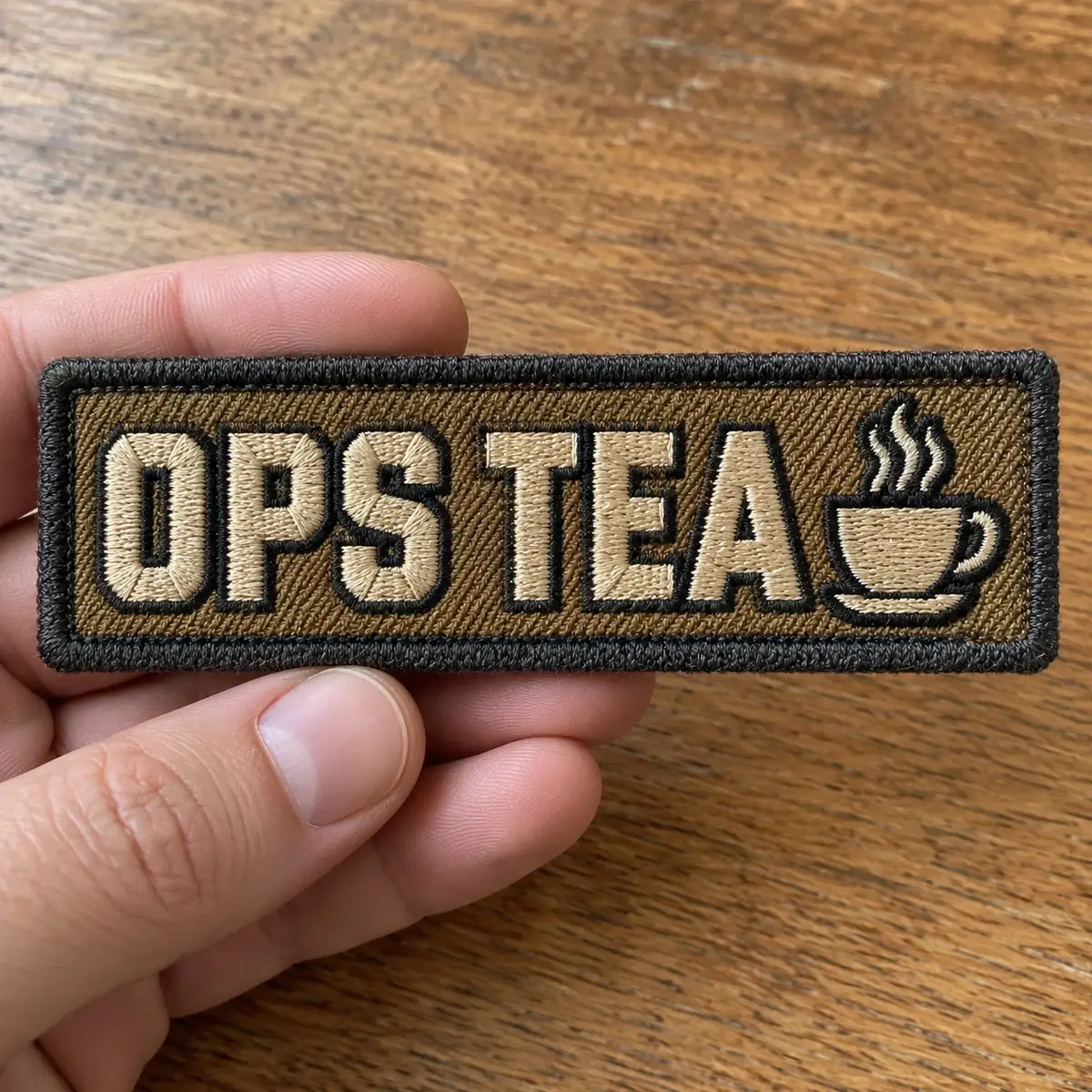Embroidered OPS TEA patch with a steaming cup, held in hand.