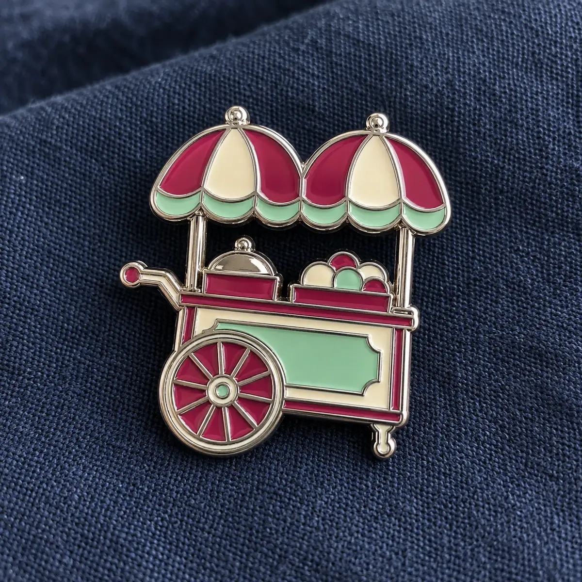 Silver enamel pin of a food cart with striped umbrellas on navy fabric.